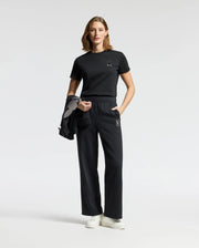A woman stands against a plain background wearing the Psycho Bunny WOMENS ALEXANDER GRAPHIC TEE and matching pants with Lurex embroidery. She holds a black jacket in one hand and pairs her outfit with white sneakers.