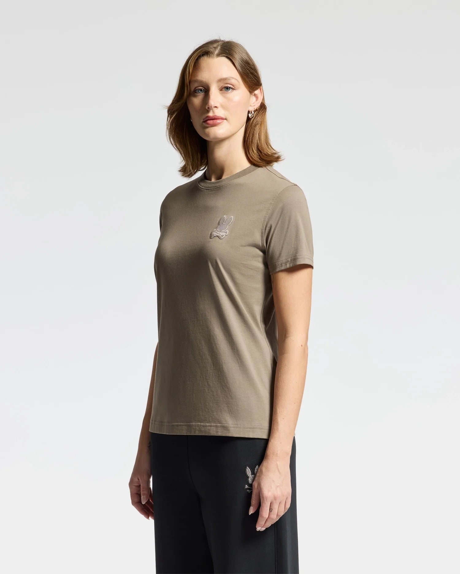 A person with shoulder-length hair is wearing the Psycho Bunny WOMENS ALEXANDER GRAPHIC TEE - B2U164E200, a brown graphic tee made from Pima cotton featuring a small logo on the chest, paired with dark pants. They are standing against a plain white background.