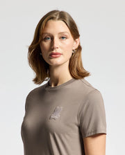 A woman with shoulder-length brown hair is wearing the gray WOMENS ALEXANDER GRAPHIC TEE - B2U164E200 by Psycho Bunny, featuring a small Lurex embroidered bunny logo on the chest. She stands against a plain white background, gazing forward.
