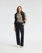 A woman stands confidently wearing a casual outfit featuring the chic WOMENS ALEXANDER GRAPHIC TEE - B2U164E200 by Psycho Bunny in brown, paired with black logo pants, a draped black hoodie, and white sneakers. The background is plain white.