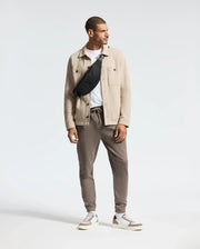 A man stands against a plain background, dressed in a beige jacket over a white shirt, complemented by gray jogger pants and white sneakers. Draped across his chest is the MENS CROSS BODY BAG - B6A181E200 by Psycho Bunny. He looks to his left with a relaxed posture.