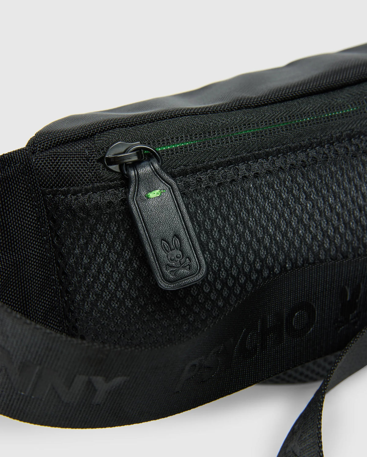 Close-up of the MENS CROSS BODY BAG - B6A181E200 by Psycho Bunny, crafted from black nylon and featuring a mesh pocket alongside a zipper pull adorned with a small bunny logo. The bag includes a strap with printed text adjacent to the pocket and boasts a subtle green accent near the zipper, combining style and functionality for everyday use.