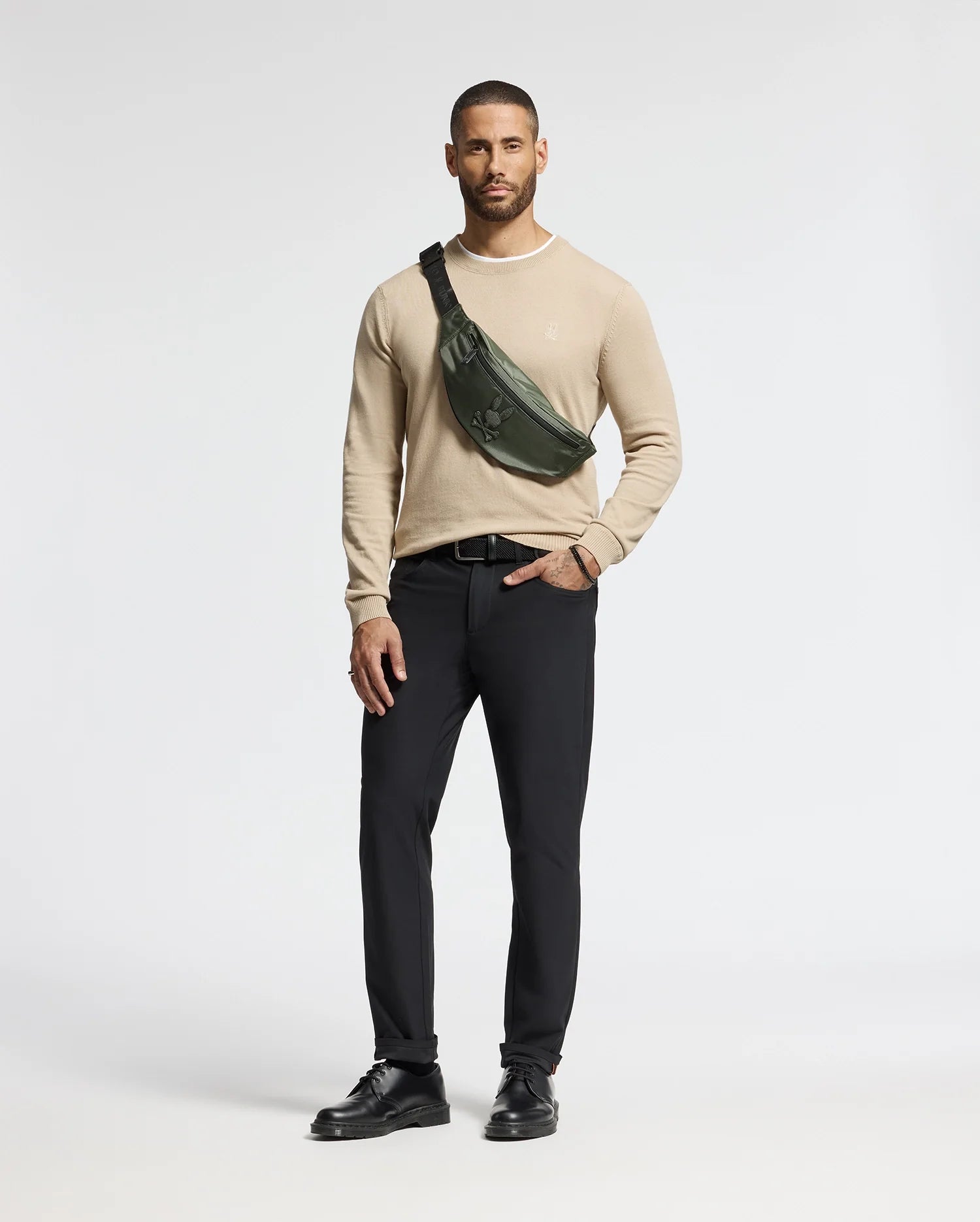 Against a white backdrop, a man wears a beige sweater, black pants, and shoes. A Psycho Bunny MENS CROSS BODY BAG - B6A181E200 in dark green is slung across his chest. With his right hand in his pocket, he exudes casual style with this chic accessory.