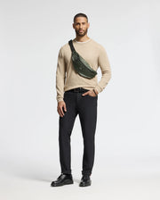 Against a white backdrop, a man wears a beige sweater, black pants, and shoes. A Psycho Bunny MENS CROSS BODY BAG - B6A181E200 in dark green is slung across his chest. With his right hand in his pocket, he exudes casual style with this chic accessory.
