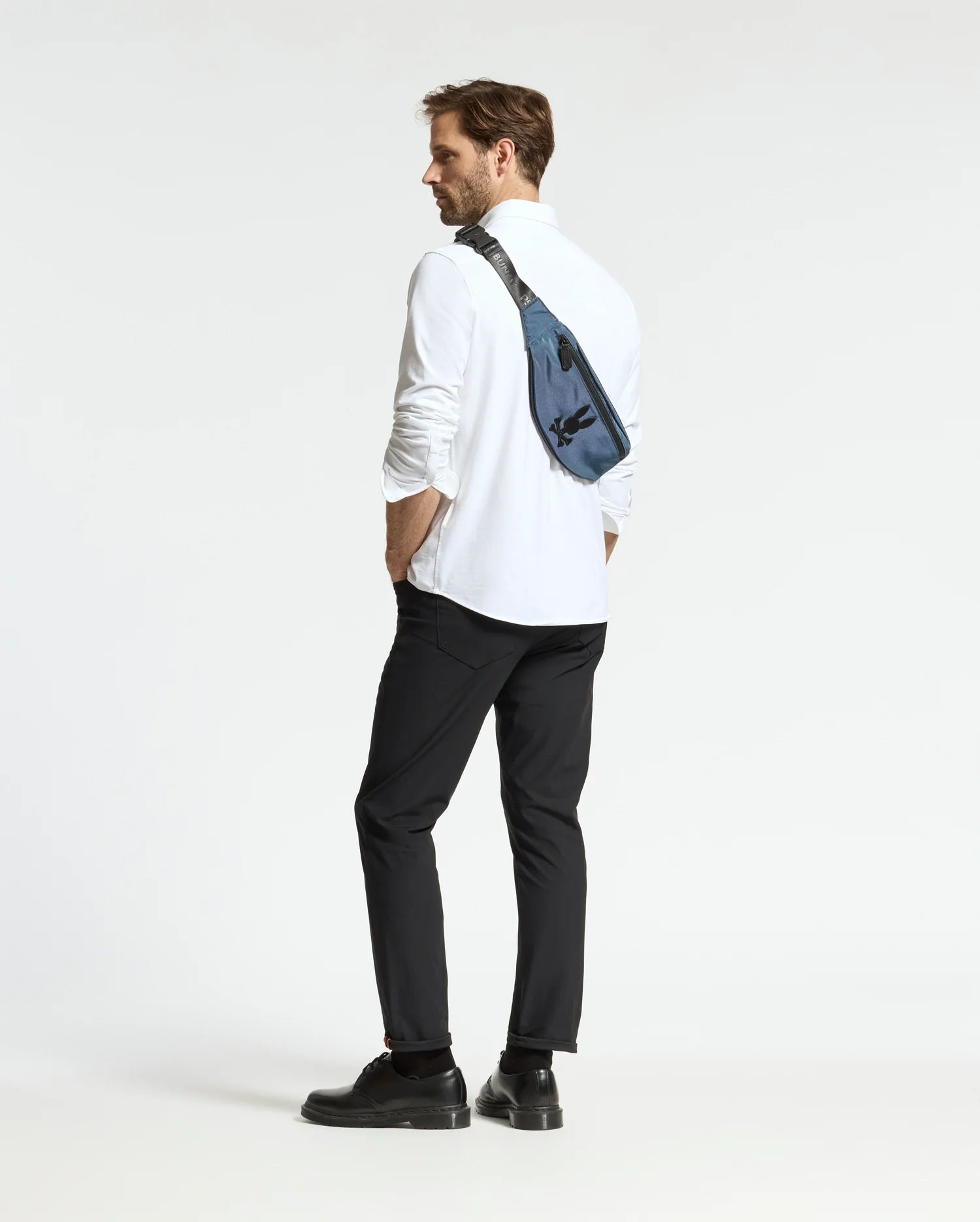 A man with a beard stands in profile, dressed in a white shirt, black pants, and black shoes. He carries the Psycho Bunny MENS CROSS BODY BAG - B6A181E200 made of durable nylon across his shoulder. The background is plain white.