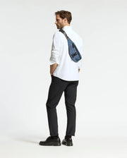 A man with a beard stands in profile, dressed in a white shirt, black pants, and black shoes. He carries the Psycho Bunny MENS CROSS BODY BAG - B6A181E200 made of durable nylon across his shoulder. The background is plain white.