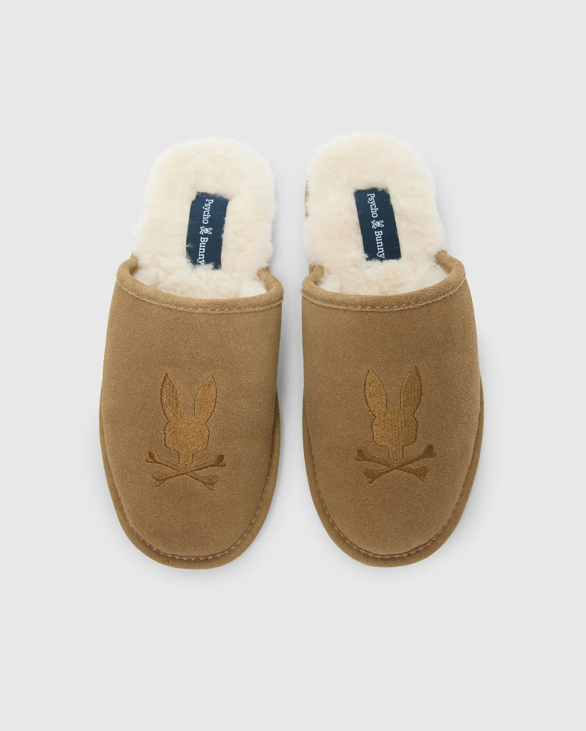 These men's suede slippers from Psycho Bunny feature a brown cow suede leather exterior and a plush white fleece lining. The embroidered rabbit skull and crossbones add a distinctive flair to the genuine shearling comfort they provide.