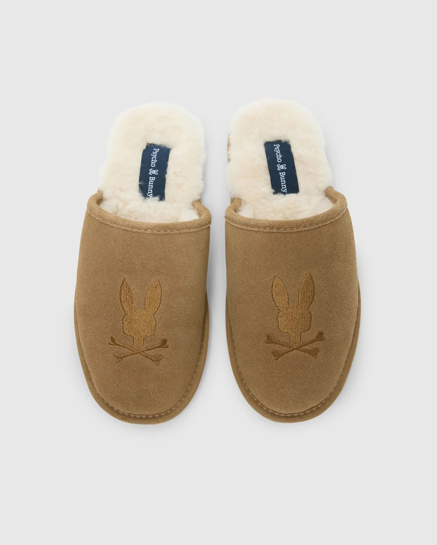 These men's suede slippers from Psycho Bunny feature a brown cow suede leather exterior and a plush white fleece lining. The embroidered rabbit skull and crossbones add a distinctive flair to the genuine shearling comfort they provide.