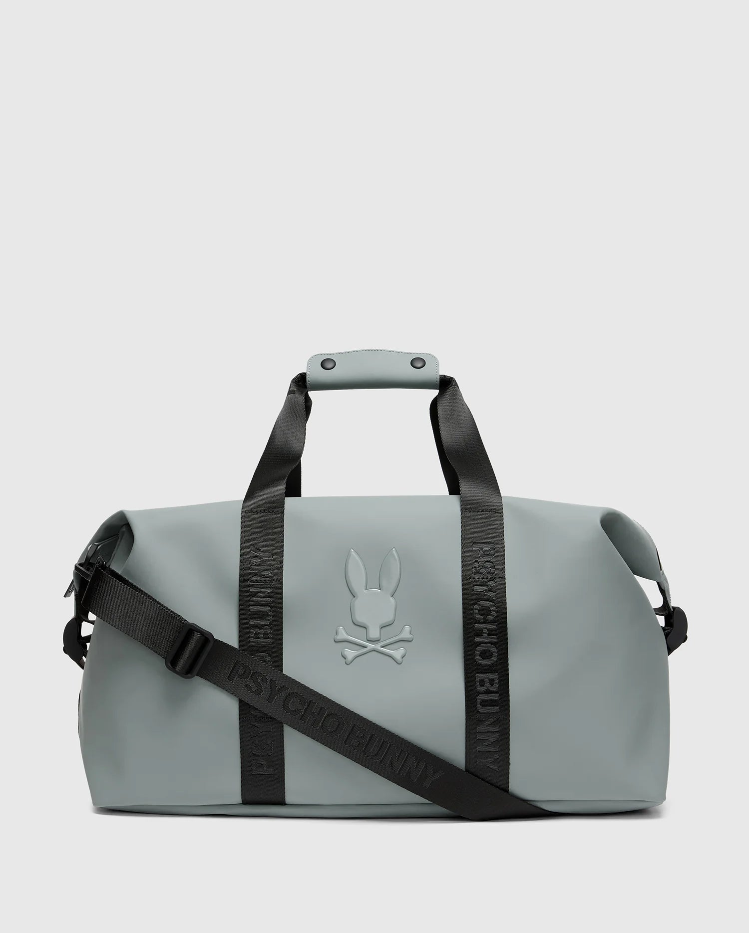 A sleek light gray men's duffle bag, model B6A472F200, from Psycho Bunny, features black handles, a detachable shoulder strap with the brand name "Psycho Bunny," and a raised bunny and crossbones logo on the front.