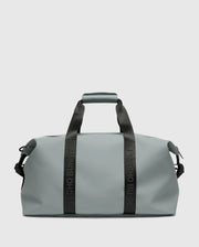 The "MENS DUFFLE BAG - B6A472F200" by Psycho Bunny is a sleek grey carry-all duffel featuring sturdy black handles and strap, the iconic logo, a minimalist design, and smooth texture. It's ideal for packing essentials for travel or gym use.