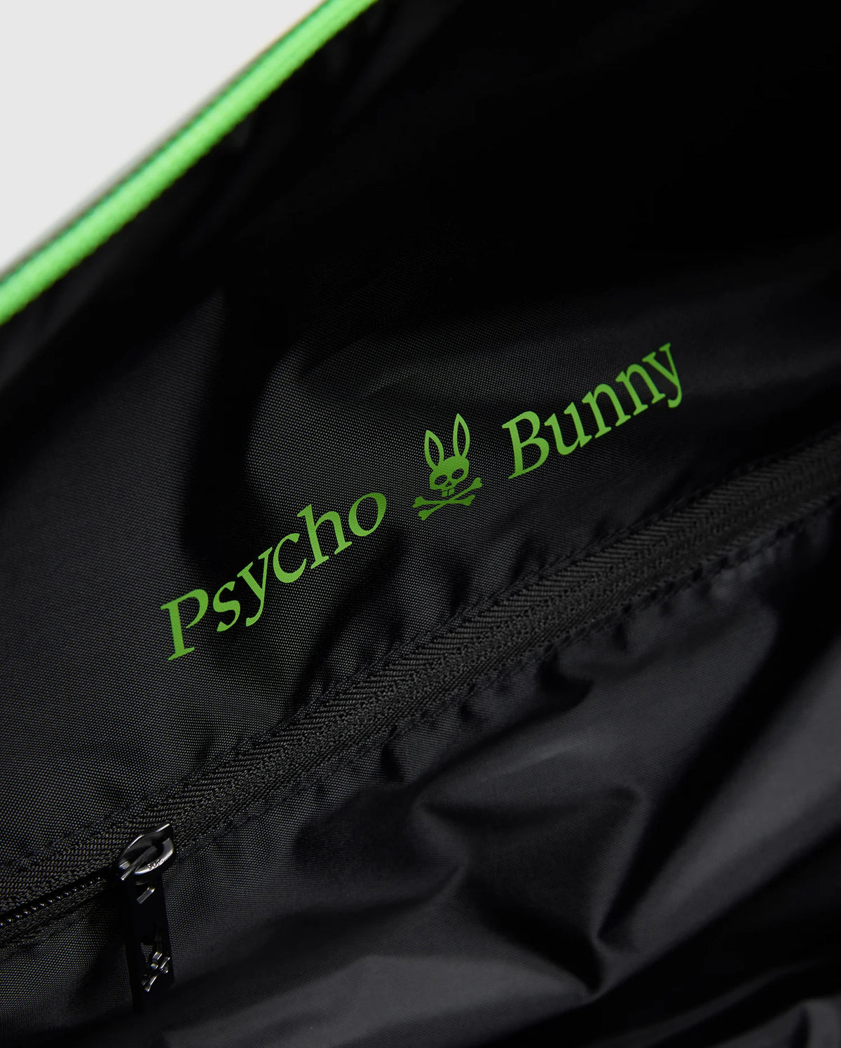 The black MENS DUFFLE BAG - B6A472F200 by Psycho Bunny features a zippered pocket inside, adorned with green "Psycho Bunny" text and a bunny skull logo. Its smooth, slightly shiny material is ideal for stylish packing.