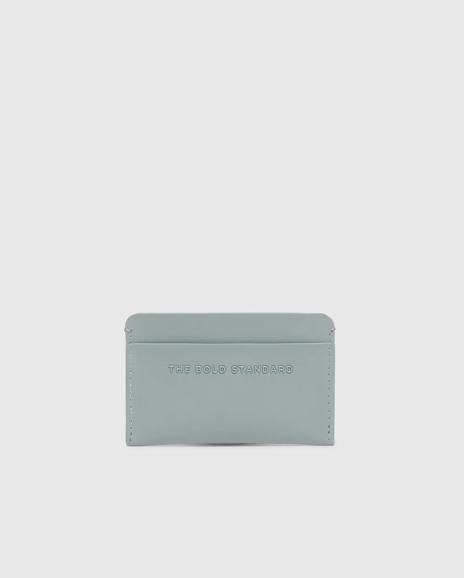 The Psycho Bunny MENS RUBBERIZED CARD CASE - B6A473F200 features "THE BOLD STANDARD" embossed on its light gray, minimalist leather design. Offering security and elegance against a white background, this piece is both stylish and functional.