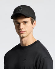 A person wearing Psycho Bunny's MENS NEW CASTLE GRAPHIC BASEBALL CAP featuring a rabbit logo, made from cotton twill with an adjustable back strap, paired with a matching black sweatshirt against a plain light gray background.