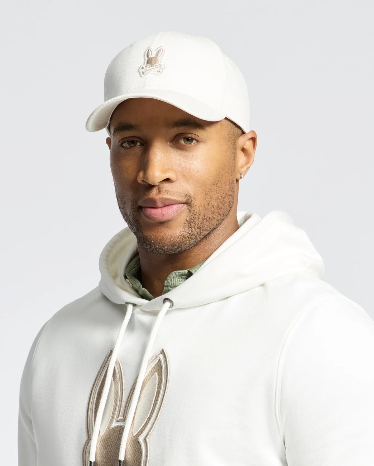 A man poses against a plain background, exuding quiet luxury in a white hoodie with a bunny design and the Psycho Bunny MENS NEW CASTLE GRAPHIC BASEBALL CAP (B6A547F200), showcasing a similar logo.