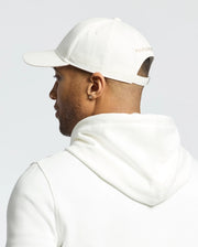 A person is shown from the back wearing a white hooded sweatshirt and the Psycho Bunny MENS NEW CASTLE GRAPHIC BASEBALL CAP, made of cotton twill with an adjustable strap and subtle side text, embodying quiet luxury against a plain, light-colored background.