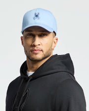 A man exuding quiet luxury wears the MENS NEW CASTLE GRAPHIC BASEBALL CAP by Psycho Bunny in light blue, paired with a black hoodie and small hoop earrings. Against a white background, his neutral expression enhances his understated style.