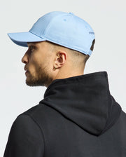 A bearded person wears an ear piercing, Psycho Bunny's Mens New Castle Graphic Baseball Cap in light blue cotton twill with subtle embroidery, and a black hoodie. They face slightly left against a plain background, radiating quiet luxury.