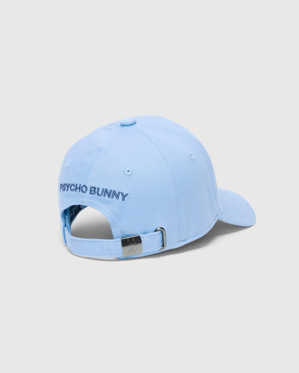 The MENS NEW CASTLE GRAPHIC BASEBALL CAP - B6A547F200 by Psycho Bunny embodies quiet luxury with PSYCHO BUNNY embroidered on the light blue cotton twill and features an adjustable strap with a metal clasp, showcasing an engraved logo against a plain gray background.