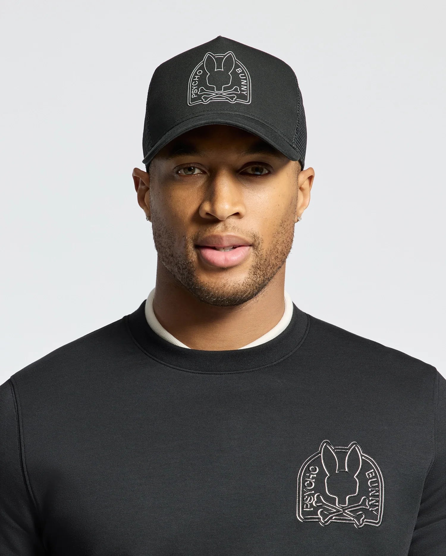 A person wearing a black sweatshirt and a Psycho Bunny MENS MELBOURNE GRAPHIC TRUCKER HAT, both featuring the brand's embroidered bunny logo. The hat's snap-back strap provides a casual touch against the plain, light-colored backdrop.