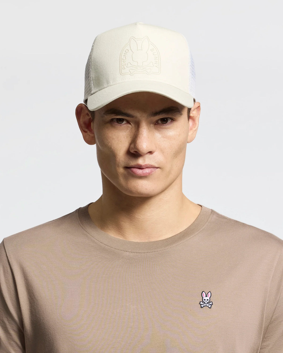 A person wears a beige Psycho Bunny MENS MELBOURNE GRAPHIC TRUCKER HAT with an embossed logo and snap-back strap, paired with a tan shirt featuring a small embroidered bunny insignia on the chest, against a plain, light-colored background.