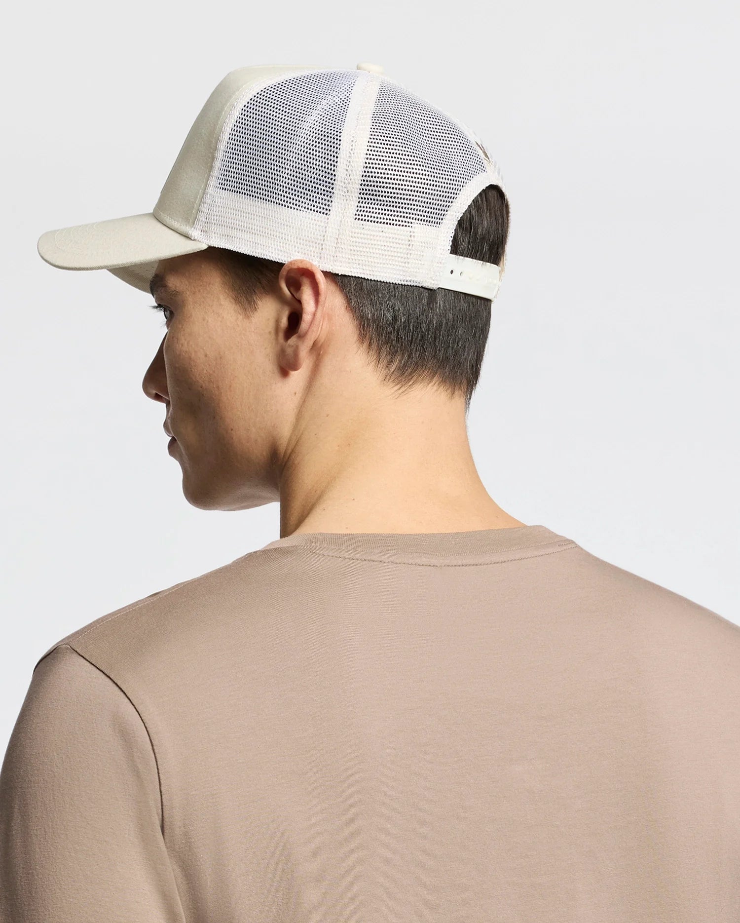 A person is seen from behind wearing the Psycho Bunny MENS MELBOURNE GRAPHIC TRUCKER HAT - B6A548F200, featuring an embroidered icon and a mesh back. The beige cap with a snap-back strap pairs seamlessly with their tan shirt against the light-colored background.