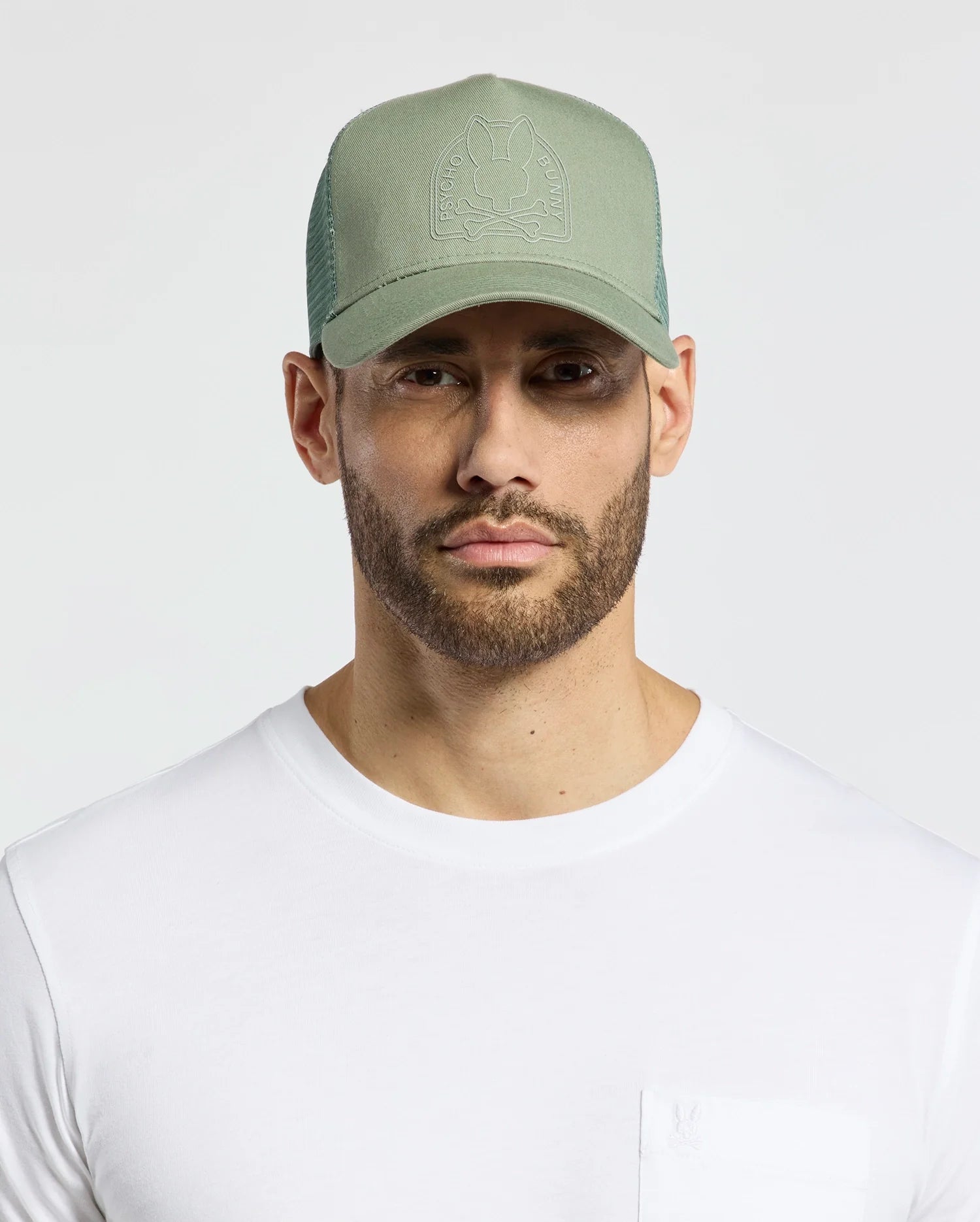 A bearded man dons a Psycho Bunny MENS MELBOURNE GRAPHIC TRUCKER HAT - B6A548F200 in light green with an embroidered icon, teamed with a plain white t-shirt for a stylish look.