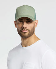 A man with a beard wears a sage green Psycho Bunny MENS MELBOURNE GRAPHIC TRUCKER HAT - B6A548F200 and a white t-shirt. The cap, featuring an embroidered icon, blends fashion and function as he stands against a light, neutral background.