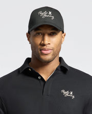 A man wears a Psycho Bunny black 100% cotton polo shirt and the MENS DESERT BASEBALL CAP (B6A549F200) with the logo, set against a plain backdrop.