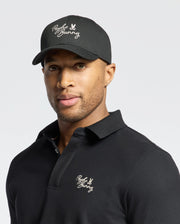 A man wearing a black Psycho Bunny MENS DESERT BASEBALL CAP - B6A549F200 and a black 100% cotton polo shirt with white embroidered text and logos stands against a plain white background.