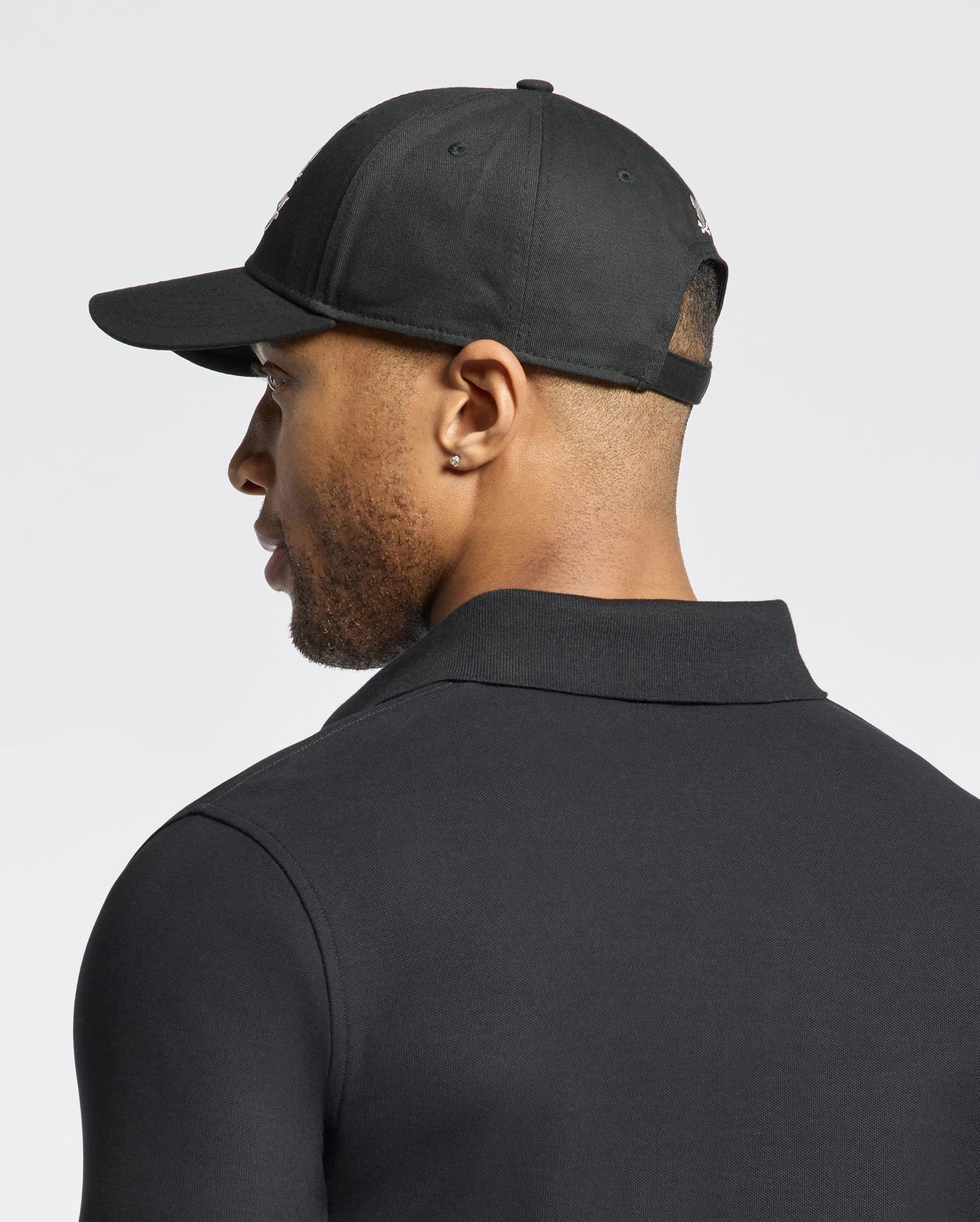 A man, embracing West Coast style, wears a Psycho Bunny MENS DESERT BASEBALL CAP - B6A549F200 in black with an adjustable strap and a small earring in his left ear. Wearing a black collared shirt, he is seen from behind against a plain light-colored background.