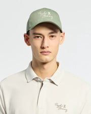 Wearing a Psycho Bunny MENS DESERT BASEBALL CAP in sage green with an embroidered wordmark and adjustable strap, paired with a light beige polo shirt featuring the same logo, they stand against a plain light background.