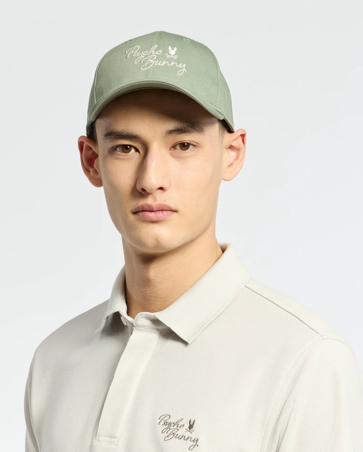 A person wears the Psycho Bunny Men's Desert Baseball Cap in light green, showcasing an embroidered wordmark and subtle logo with an adjustable strap. They pair it with a light gray polo shirt featuring matching embroidery, set against a plain, light-colored background.