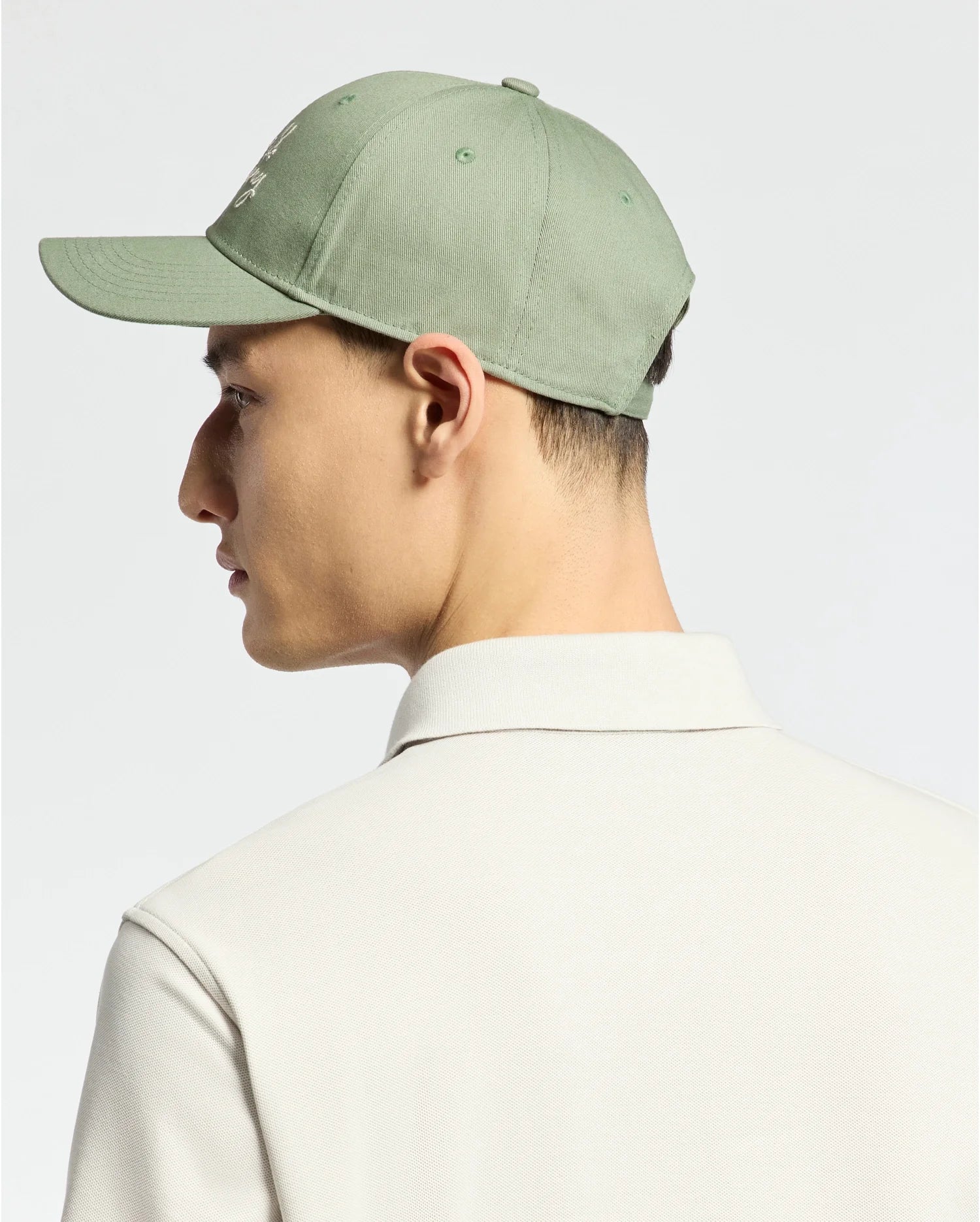 Someone is wearing a MENS DESERT BASEBALL CAP - B6A549F200 by Psycho Bunny, featuring an embroidered wordmark and adjustable strap, in light green cotton. They're facing sideways in a white collared shirt against a neutral background.