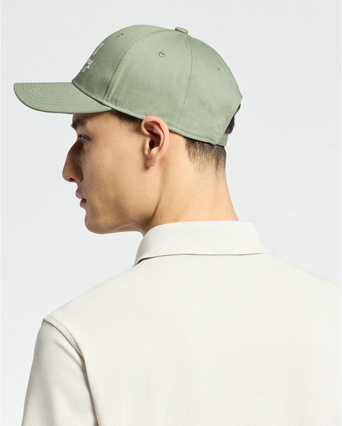 Someone is wearing a MENS DESERT BASEBALL CAP - B6A549F200 by Psycho Bunny, featuring an embroidered wordmark and adjustable strap, in light green cotton. They're facing sideways in a white collared shirt against a neutral background.