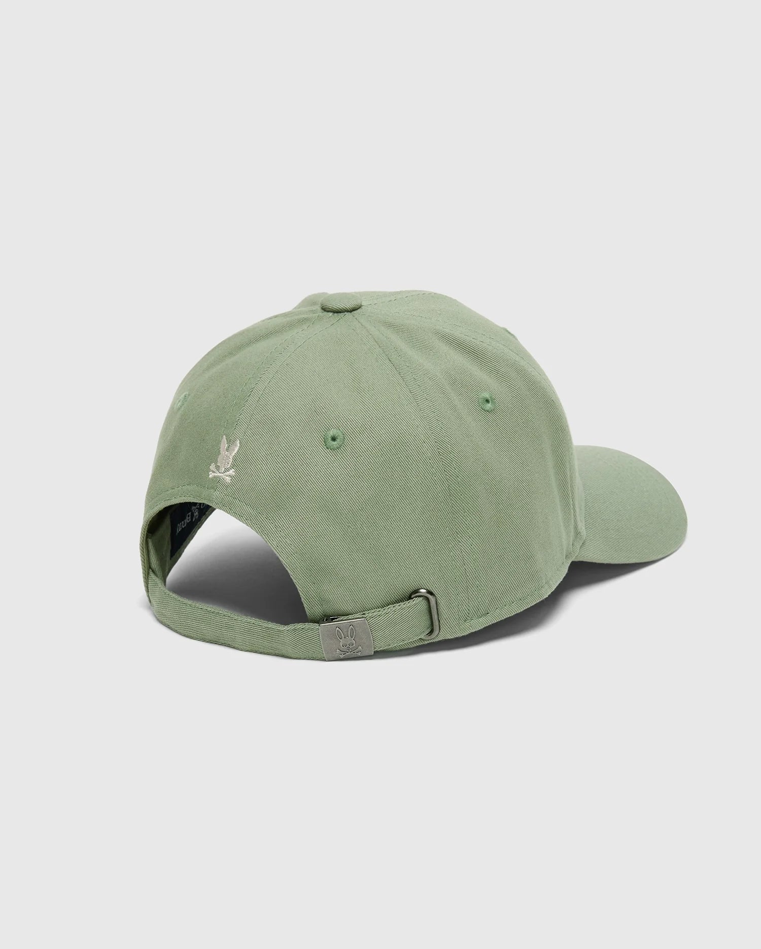 The Psycho Bunny MENS DESERT BASEBALL CAP - B6A549F200 is shown from the back, displaying a light green cotton material with an adjustable metal buckle strap. A small embroidered wordmark appears above the opening, all set against a plain gray background.