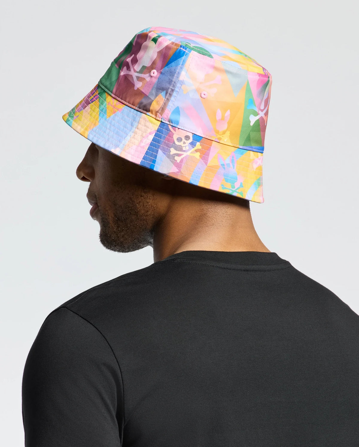 Wearing a Psycho Bunny MENS MILOS BUCKET REVERSIBLE HAT, detailed with skulls and rabbits, the person enhances their black shirt against a plain backdrop.