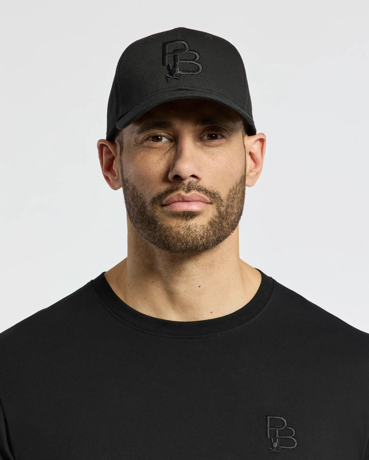 A bearded man wears a black Psycho Bunny MENS NICHOLSON PB BUNNY GRAPHIC BASEBALL CAP with an adjustable strap, showcasing a PB-embroidered design, and pairs it with a black shirt featuring subtle logos. His expression is neutral against a plain background.