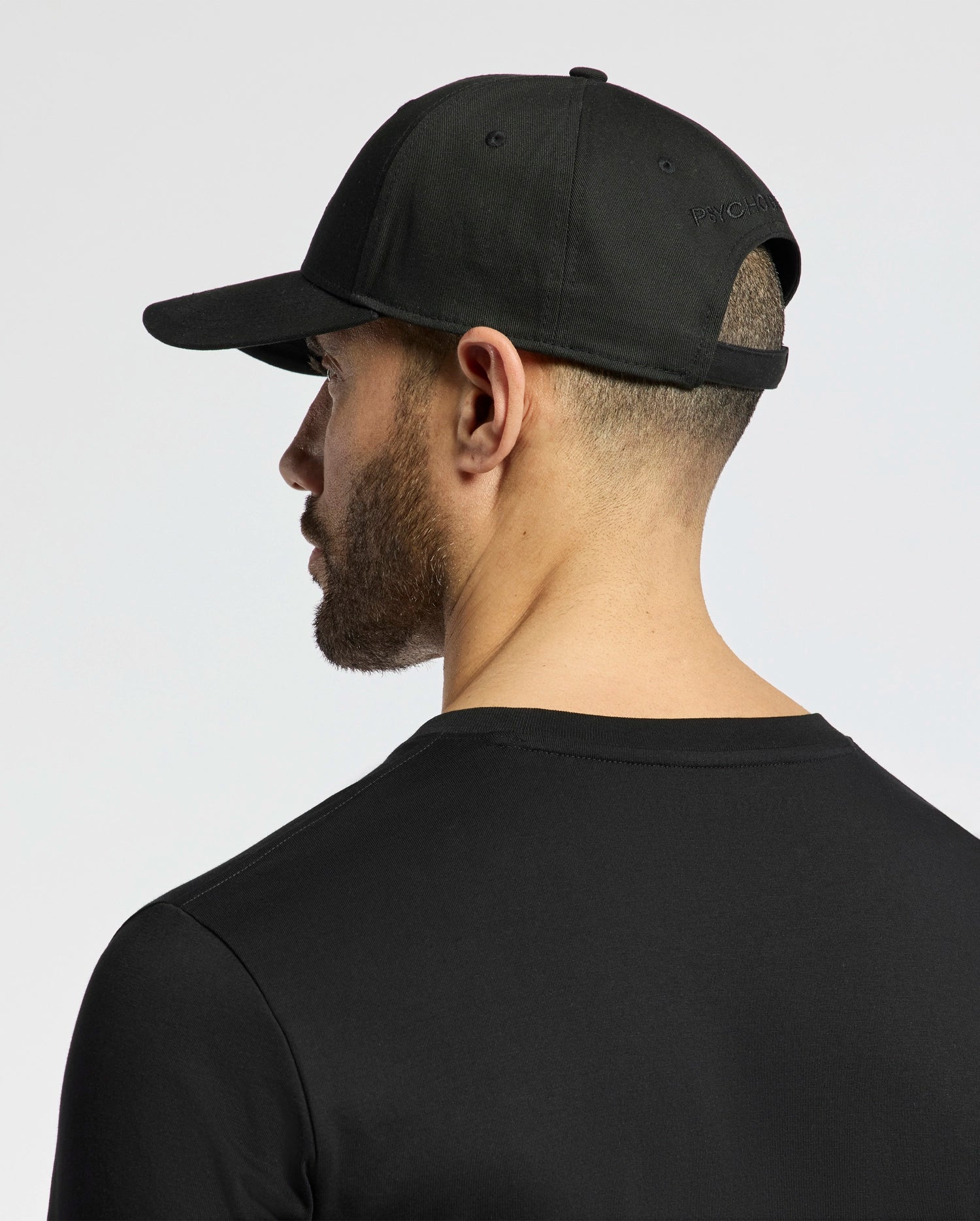 A man, seen from behind and slightly to the side against a plain backdrop, is wearing a Psycho Bunny MENS NICHOLSON PB BUNNY GRAPHIC BASEBALL CAP - B6A553F200 in black cotton twill with an adjustable backstrap. The PB-embroidered design enhances the cap's stylish appeal.