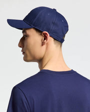 A person wearing a Psycho Bunny navy blue MENS NICHOLSON PB BUNNY GRAPHIC BASEBALL CAP (B6A553F200) with an adjustable backstrap and a matching t-shirt is shown from the side. The cap's strap subtly reflects light, hinting at embroidery. The background is plain and light-colored.