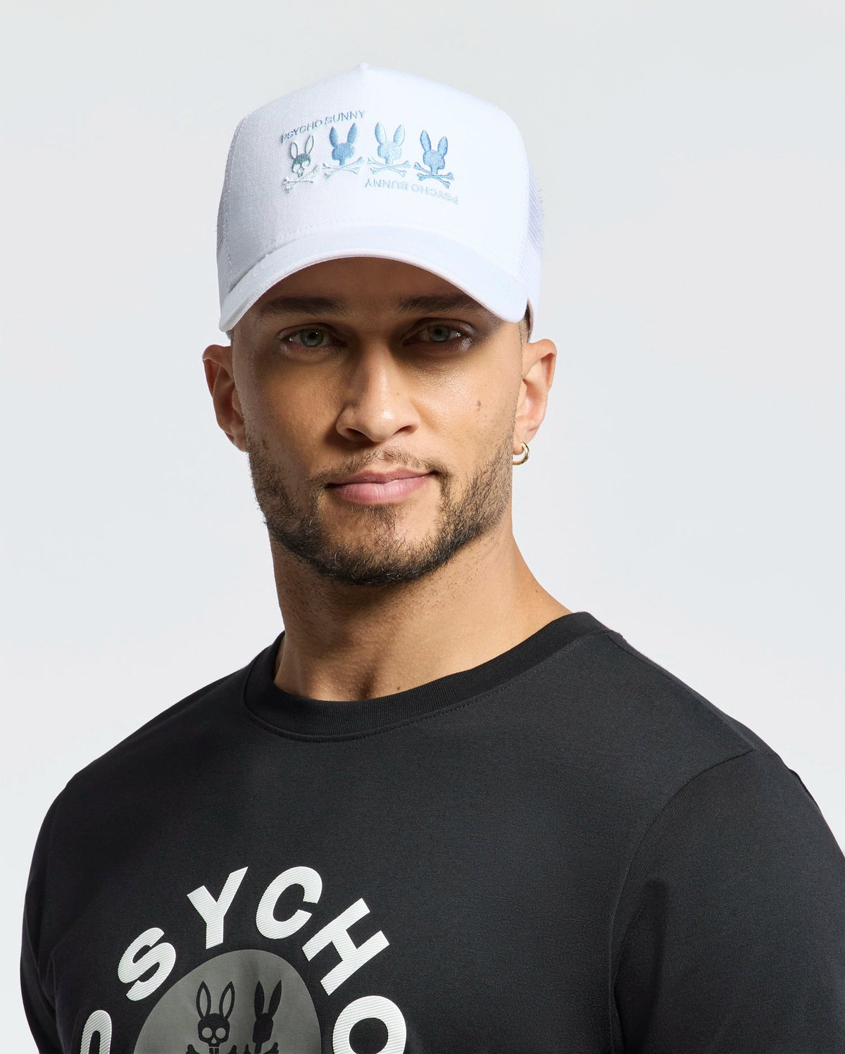 A man in a white Psycho Bunny Men's Oceania Embroidered Graphic Trucker Hat featuring cartoon mice and text complements his streetwear vibe with a black shirt adorned with stylized lettering. A small earring enhances his neutral look against the light background.