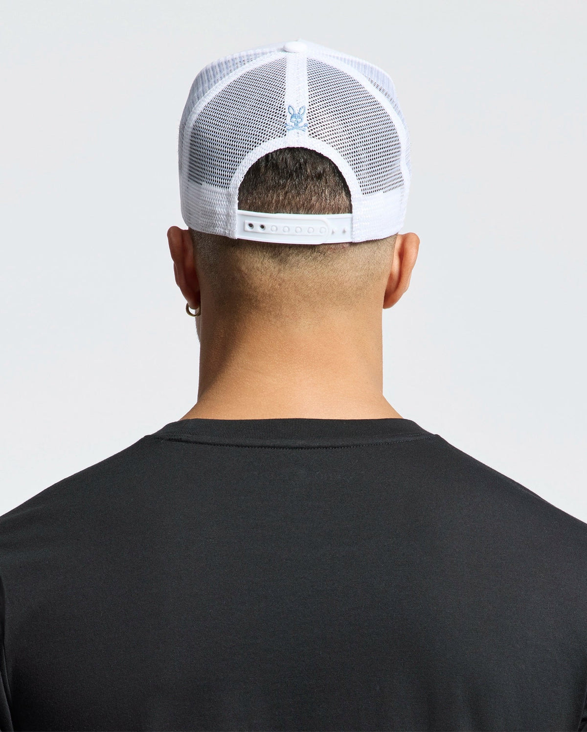 A person with short hair, facing away from the camera, wears a Psycho Bunny MENS OCEANIA EMBROIDERED GRAPHIC TRUCKER HAT in white mesh and a black shirt. The cap has an adjustable strap and embroidered bunny graphic on the back, offering a streetwear vibe against a plain light background.