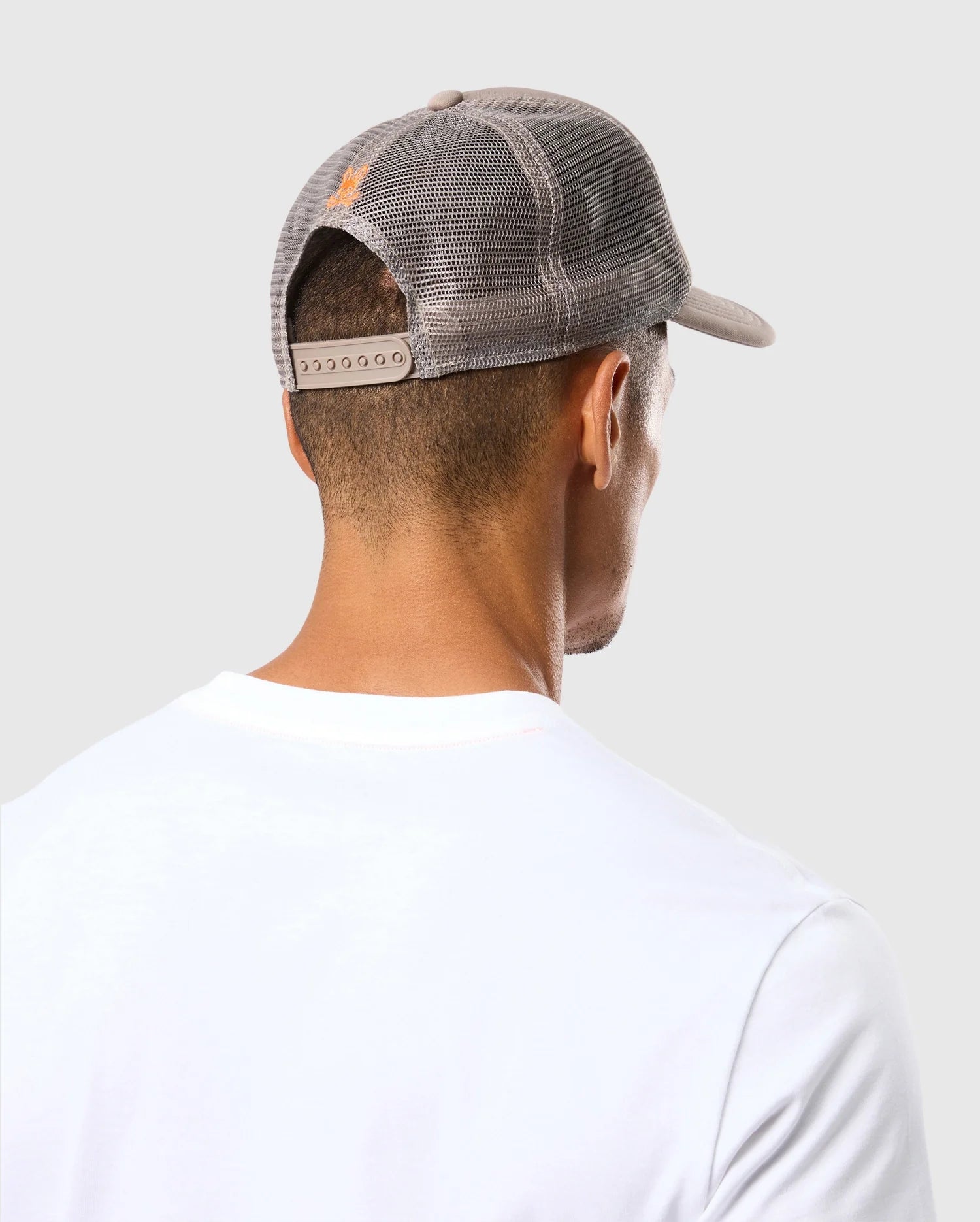 A person is shown from behind wearing a white shirt and the Psycho Bunny MENS PARIS TRUCKER CAP - B6A591C200, which features mesh paneling. The cap is gray and showcases an orange bunny logo on the back. The scene is set against a plain light gray background, and the person's head is slightly tilted to the right.