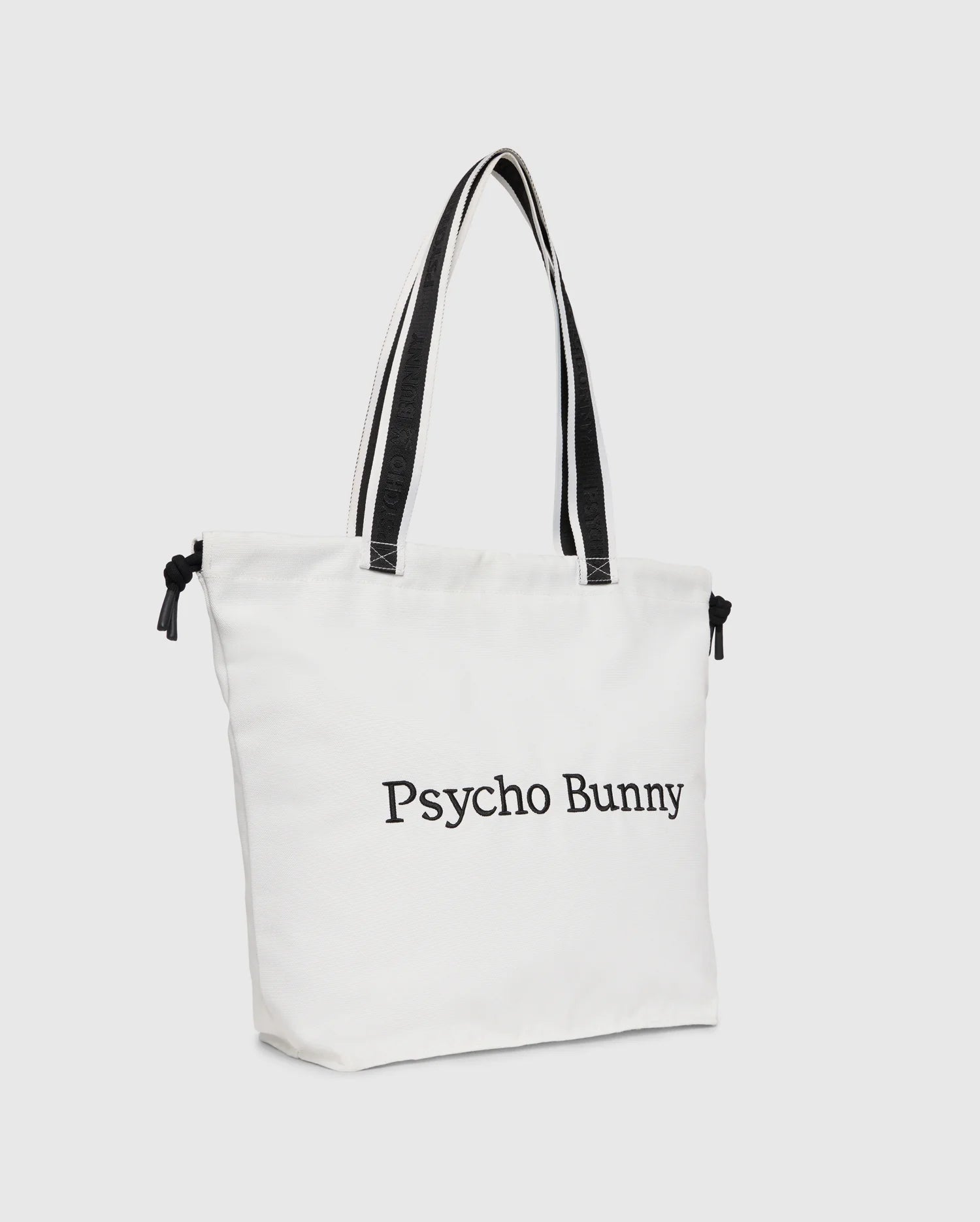 The "TENNIS TOTE - B6A596C200" by Psycho Bunny is a white canvas tote bag with black handles, featuring the text "Psycho Bunny" on the front and an embroidered logo for an elegant touch.
