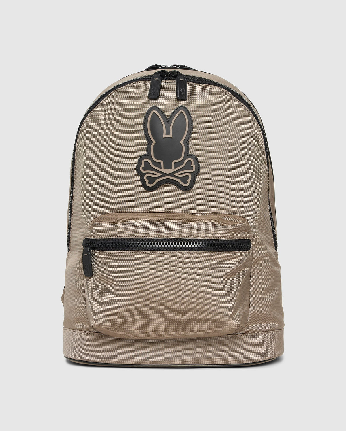 A beige Psycho Bunny MENS DOME BACKPACK - B6A628Z1BP with a black zipper, front pocket, and black emblem of a rabbit head with crossed bones. The minimalist design features a padded laptop compartment and two zipper pulls at the top for added style.