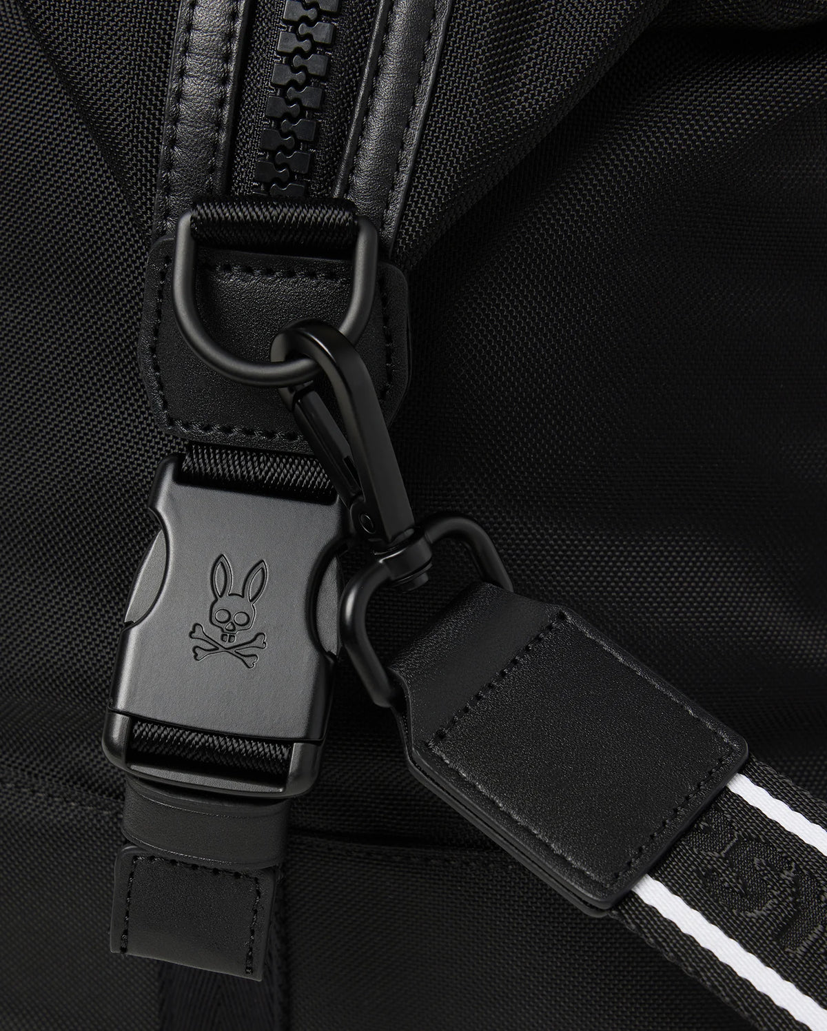 Close-up image of a backpack strap featuring a black buckle with a bunny and crossbones logo engraved on it. The strap includes a black carabiner clip and a white-striped detail to enhance its design, making it perfect for pairing with your Psycho Bunny MENS WEEKENDER BAG - B6A629Z1BG or travel luggage. The fabric is black and textured.
