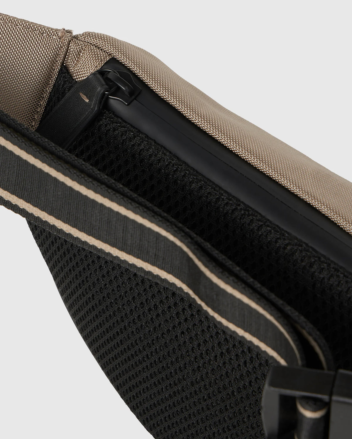 Close-up image of a beige and black sling bag. The Psycho Bunny MENS BELT BAG - B6A631Z1BP features a zipper closure, rubber bunny patch, black mesh back panel for breathability, and an adjustable strap with beige accents. The materials appear durable and well-constructed.