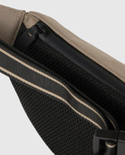 Close-up image of a beige and black sling bag. The Psycho Bunny MENS BELT BAG - B6A631Z1BP features a zipper closure, rubber bunny patch, black mesh back panel for breathability, and an adjustable strap with beige accents. The materials appear durable and well-constructed.