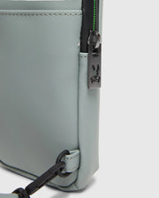 Close-up of the corner of a light gray Psycho Bunny MENS RUBBERIZED CROSSBODY (B6A635F200) crafted from durable polyester, featuring a black zipper with a sleek rabbit logo, visible stitching, and an attached black crossbody strap.