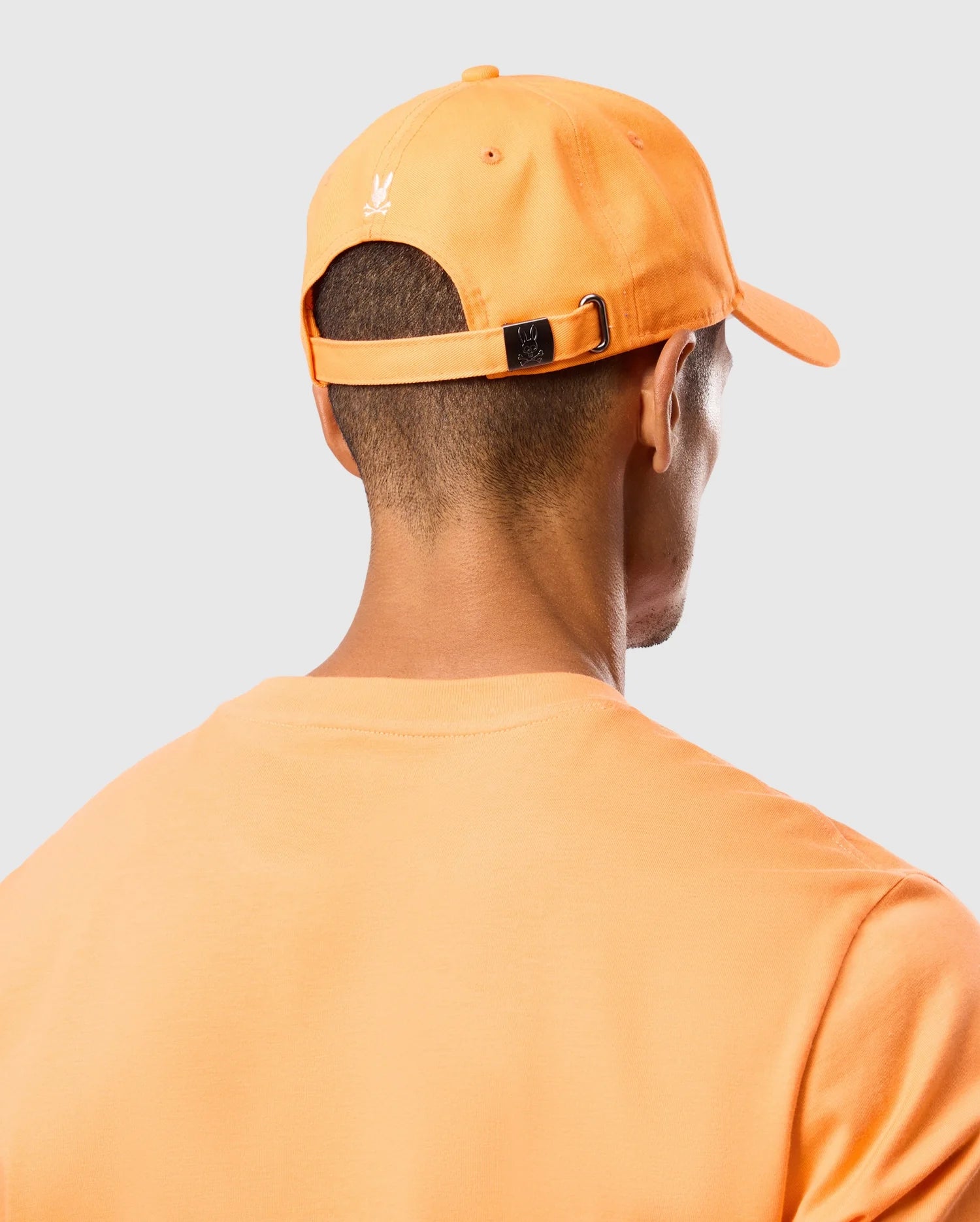 The image shows the back of a person wearing an orange Psycho Bunny MENS KAYDEN BASEBALL CAP - B6A677C200 and an orange T-shirt. The person's head is turned slightly to the right, and the cap features a Bunny logo and has an adjustable strap with a metal buckle at the back. The background is plain and light-colored.