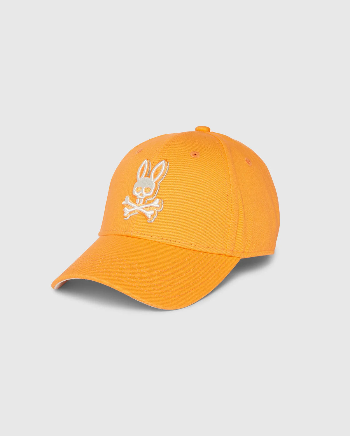 The Psycho Bunny MENS KAYDEN BASEBALL CAP - B6A677C200 is an orange cotton twill baseball cap with a white embroidered bunny logo featuring a head above two crossed bones on the front. It also includes a curved brim and an adjustable strap at the back.